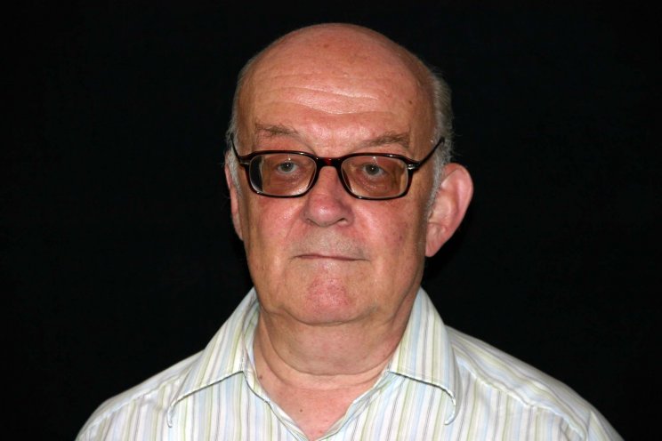 Das Porträt-Foto zeigt einen älteren Mann. Er hat größtenteils eine Glatze und nur an den Seiten graue Haare. Er trägt eine Brille sowie ein weiß gestreiftes Hemd. Das Foto ist vor einem schwarzen Hintergrund aufgenommen.