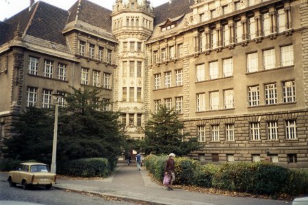 Das Foto zeigt ein Gebäude mit einem auffälligen Turm in der Mitte. Es hat mehrere Stockwerke mit vielen Fenstern und ornamentalen Details an der beigen Fassade. Vor dem Haupteingang befinden sich Grünanlagen mit Hecken und Nadelbäumen. Auf der davor liegenden Straße steht im linken Bildrand ein gelber Trabant. Vor dem Gebäude sind einige Personen zu sehen, die auf dem Gehweg entlanggehen. Eine ältere Person mit einer Tasche ist im Vordergrund erkennbar.