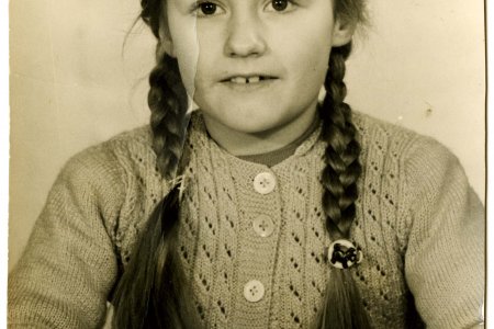 Die Schwarz-Weiß-Fotografie zeigt ein Mädchen. Es trägt eine Strickjacke mit einem Muster und Knöpfen. Die Haare sind zu zwei langen Zöpfen geflochten, die über die Schultern nach vorne fallen. Das Mädchen sitz an einem Tisch und hat die Hände gefaltet.