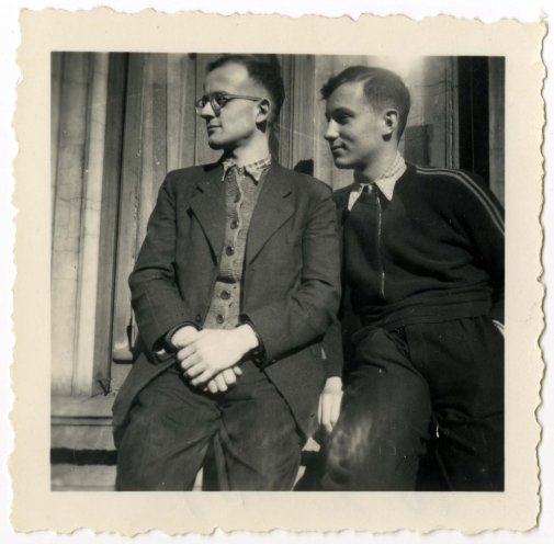 Die Schwarz-Weiß-Fotografie zeigt zwei junge Männer. Sie lehnen an einer Gebäudefassade. Der Mann links im Bild trägt eine Brille und eine Anzugjacke mit einer zugeknöpften Strickjacke darunter, der Mann rechts im Bild trägt einen dunklen Pullover.