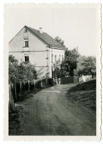 Die Schwarz-Weiß-Fotografie zeigt ein Haus an einer unbefestigten Straße. Das zweistöckige Haus hat mehrere Fenster und ist aus hellen Materialien gebaut. Es hat dekorative Ecksteine und ein steiles Pultdach mit einem Schornstein und einem kleinen runden Fenster an der Spitze. Das Grundstück wird von einem Zaun begrenzt, der sich entlang der Straße fortsetzt. Entlang der Zaunlinie wachsen Bäume und Sträucher.