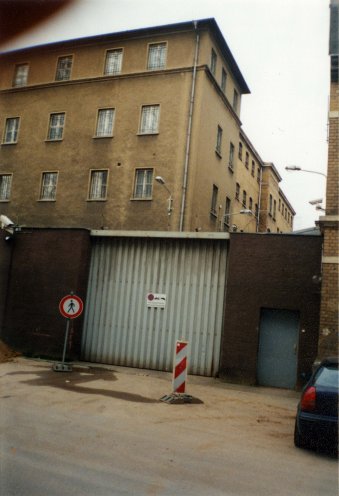 Das Foto zeigt Teile eines mehrstöckigen Gebäudes mit einer hellbraunen Fassade. Es verfügt über Fensterreihen mit Sicherheitsstäben. An den Gebäudeaußenwänden sind etliche Straßenlampen angebracht. Vor dem Gebäude befindet sich ein ummauerter Bereich mit einem großen Wellblechtor sowie einer grauen Tür. Am Tor ist ein Schild mit einem Halteverbotshinweis angebracht. Vor dem Tor steht ein Schild, das auf das Verbot von Fußgängern hinweist. Daneben steht ein rot-weiß gestreifter Absperrpfosten. Am linken Bildrand ist eine Überwachungskamera zu sehen. Im Vordergrund unten rechts ist ein Teil eines blauen Autos zu sehen.
