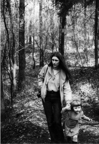 Das Schwarzweißfoto zeigt eine junge Frau mit einem kleinen Kind in einem Wald. Die Frau trägt eine helle Jacke über einem hellen Oberteil, dunkle Hosen und hat langes, glattes Haar. Sie hält die Hand des Kindes, das ein Kleid trägt und einen kurzen Bobhaarschnitt mit Pony hat. Im Hintergrund ist ein Wald mit Bäumen und Geäst zu sehen. Der Waldboden ist mit Laub und Unterholz bedeckt.