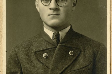 Das Porträtfoto zeigt einen jungen Mann. Er trägt eine runde Brille mit dünnem Rahmen, Einen Mantel mit breitem Revers und zwei deutlich sichtbaren Knöpfen, eine Krawatte und ein Hemd darunter sowie eine nach hinten gekämmte Frisur. Der Mann blickt direkt in die Kamera.