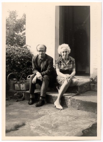 Dieses Schwarz-Weiß-Foto zeigt eine ältere Frau und einen mittelalten Mann sitzend auf einer Steinstufe vor der Türschwelle eines Hauses. Die Frau hat helles, kurzes Haar und trägt eine Brille sowie ein gemustertes kurzärmeliges Oberteil und einen Rock. Der Mann trägt eine dunkle Anzugjacke und Hose. Er trägt ebenfalls eine Brille und hat einen zurückweichenden Haaransatz. Auf der linken Seite des Bildes sind Sträucher und eine Gießkanne zu sehen.