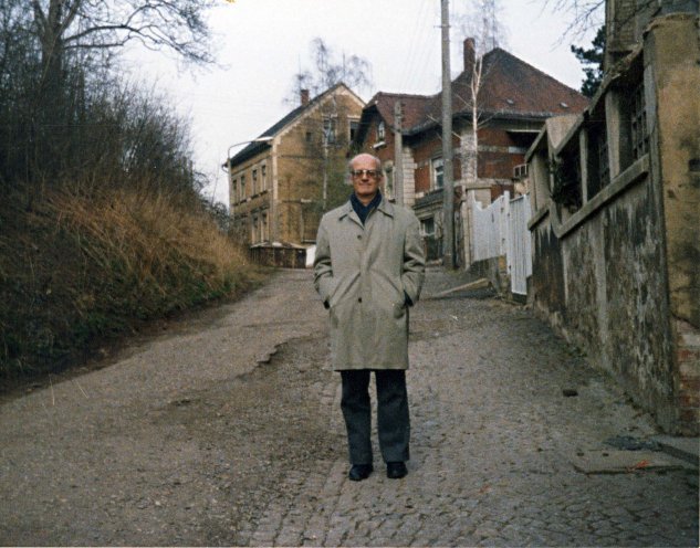In der Mitte des Fotos steht ein Mann auf einer kleinen Straße. Die Straße selbst ist uneben und besteht aus Schotter und Pflastersteinen. Der Mann trägt einen hellen Trenchcoat, eine dunkle Hose und eine Brille. Er hat die Hände in den Manteltaschen. Auf der rechten Seite der Straße stehen ältere, teilweise zerfallene Gebäude mit Ziegel- und Steinelementen. Die linke Seite des Bildes zeigt einen überwucherten Hang und kahle Bäume.