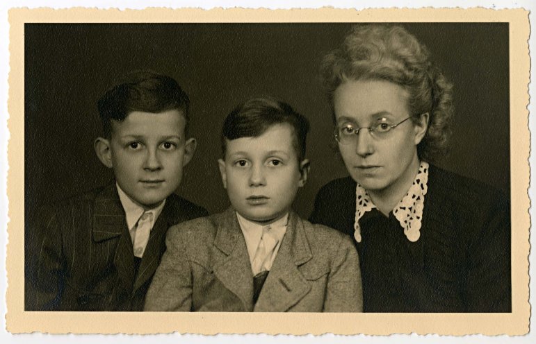 Das Schwarz-Weiß-Porträtfoto zeigt zwei Jungen und eine Frau. Der links sitzende Junge trägt eine helle Anzugjacke, der Junge in der Mitte trägt eine dunkle Jacke, beide haben einen ähnlichen gekämmten Kurzhaarschnitt und tragen weiße Hemden. Die Frau sitzt rechts und trägt eine Brille. Ihr Haar ist hochgesteckt und sie trägt einen mit Mustern verzierten weißen Kragen zu ihrer dunklen Kleidung.