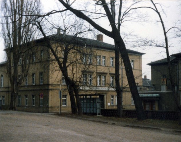 Das Bild zeigt ein dreistöckiges beiges Gebäude. Es hat eine symmetrische Fassade mit mehreren Fenstern und einem zentralen Eingang mit blauen Türen. Über dem Eingang steht Erweiterte Oberschule. Die Bäume im Vordergrund tragen keine Blätter und rahmen den Blick auf das Gebäude ein. Davor befindet sich eine gepflasterte Straße. Auf der rechten Seite ist teilweise ein weiteres Gebäude sichtbar.