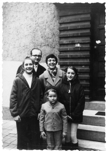 Das Schwarzweißfoto zeigt eine Gruppe von fünf Personen, die vor dem Eingang eines Gebäudes steht. Ein erwachsener Mann mit Brille in einem dunklen Anzug und Krawatte steht in der hinteren Reihe. Eine erwachsene Frau mit kurzen dunklen Haaren in einem helleren Mantel steht rechts vom Mann. Zwei junge Mädchen in dunklen Jacken stehen eines links vor dem Mann und eines rechts von der Frau. Im Vordergrund steht ein kleiner Junge in einem hellen Mantel mit Knöpfen
Die Gruppe steht vor einer teilweise mit Ziegelsteinen verkleideten Wand mit einer Tür auf einem gepflasterten Boden.