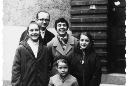 Das Schwarzweißfoto zeigt eine Gruppe von fünf Personen, die vor dem Eingang eines Gebäudes steht. Ein erwachsener Mann mit Brille in einem dunklen Anzug und Krawatte steht in der hinteren Reihe. Eine erwachsene Frau mit kurzen dunklen Haaren in einem helleren Mantel steht rechts vom Mann. Zwei junge Mädchen in dunklen Jacken stehen eines links vor dem Mann und eines rechts von der Frau. Im Vordergrund steht ein kleiner Junge in einem hellen Mantel mit Knöpfen
Die Gruppe steht vor einer teilweise mit Ziegelsteinen verkleideten Wand mit einer Tür auf einem gepflasterten Boden.