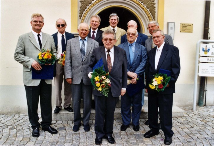 Das Bild zeigt eine Gruppe von 10 älteren Männern in Anzügen und Krawatten, die vor einem Gebäude mit einem verschnörkelten Bogenportal mit Stuckverzierungen und goldenen Akzenten stehen. Sie sind für ein formelles Foto in zwei Reihen auf einem Kopfsteinpflaster angeordnet. Mehrere der Männer halten Blumensträuße mit blauen Mappen in den Händen. Einige der Männer tragen Brillen, und ein Mann in der hinteren Reihe trägt eine Sonnenbrille.