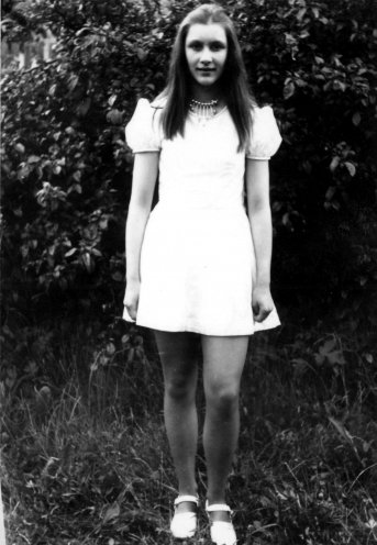 Die Schwarzweißfotografie zeigt ein Mädchen, das vor einem Busch auf einer Wiese steht. Das Mädchen trägt ein kurzes, weißes Kleid mit Puffärmeln und weiße Schuhe. Um den Hals ist eine Halskette zu sehen. Das Mädchen hat langes, glattes Haar und schaut direkt in die Kamera.